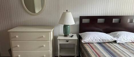 A bedroom with a white dresser, a bedside table with a lamp, a bed with white pillows, and a mirror on the wall.