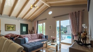 A modern living room with a sofa, ottoman, and glass coffee table. There is a door with a green frame, a painting on the wall, and a view of a pool through large windows. - Villa Jewel in Corfu (Kontokali)