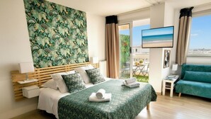 A hotel room with a bed, a green sofa, a TV, and a balcony with a view of the beach. - Furnished Studio 2** - On the edge of the Grande Plage - Swimming pool - Terrace (Biarritz)