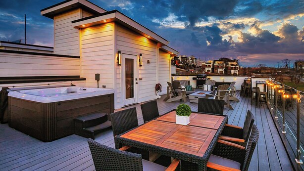 A modern outdoor deck with a hot tub, wooden tables, and chairs, overlooking a cityscape at dusk. - ROOFTOP HOT TUB- FIRE PIT- MASSIVE CITY VIEWS (Nashville)