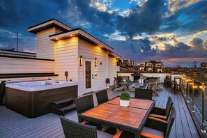 A modern outdoor deck with a hot tub, wooden tables, and chairs, overlooking a cityscape at dusk. - ROOFTOP HOT TUB- FIRE PIT- MASSIVE CITY VIEWS (Nashville)
