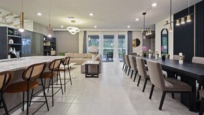 A modern kitchen with a central island, bar stools, and a dining area with a long table and chairs. - Luxury Disney Home W/private Pool Spa Game Bar (Davenport)