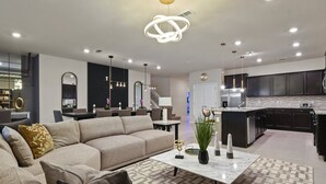 A modern living room with a beige sofa, a marble coffee table, and a kitchen area with dark cabinetry and a white countertop. - Luxury Disney Home W/private Pool Spa Game Bar (Davenport)