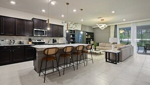 A modern kitchen with dark cabinets, a central island with bar stools, and a dining area with a sofa and table. - Luxury Disney Home W/private Pool Spa Game Bar (Davenport)