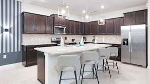 A modern kitchen with dark wood cabinets, a stainless steel refrigerator, a built-in oven, and a granite countertop. There are three bar stools and pendant lights above the island. - Windsor Island Resort 10br Villa Pool Near Disney 3063 (Davenport)