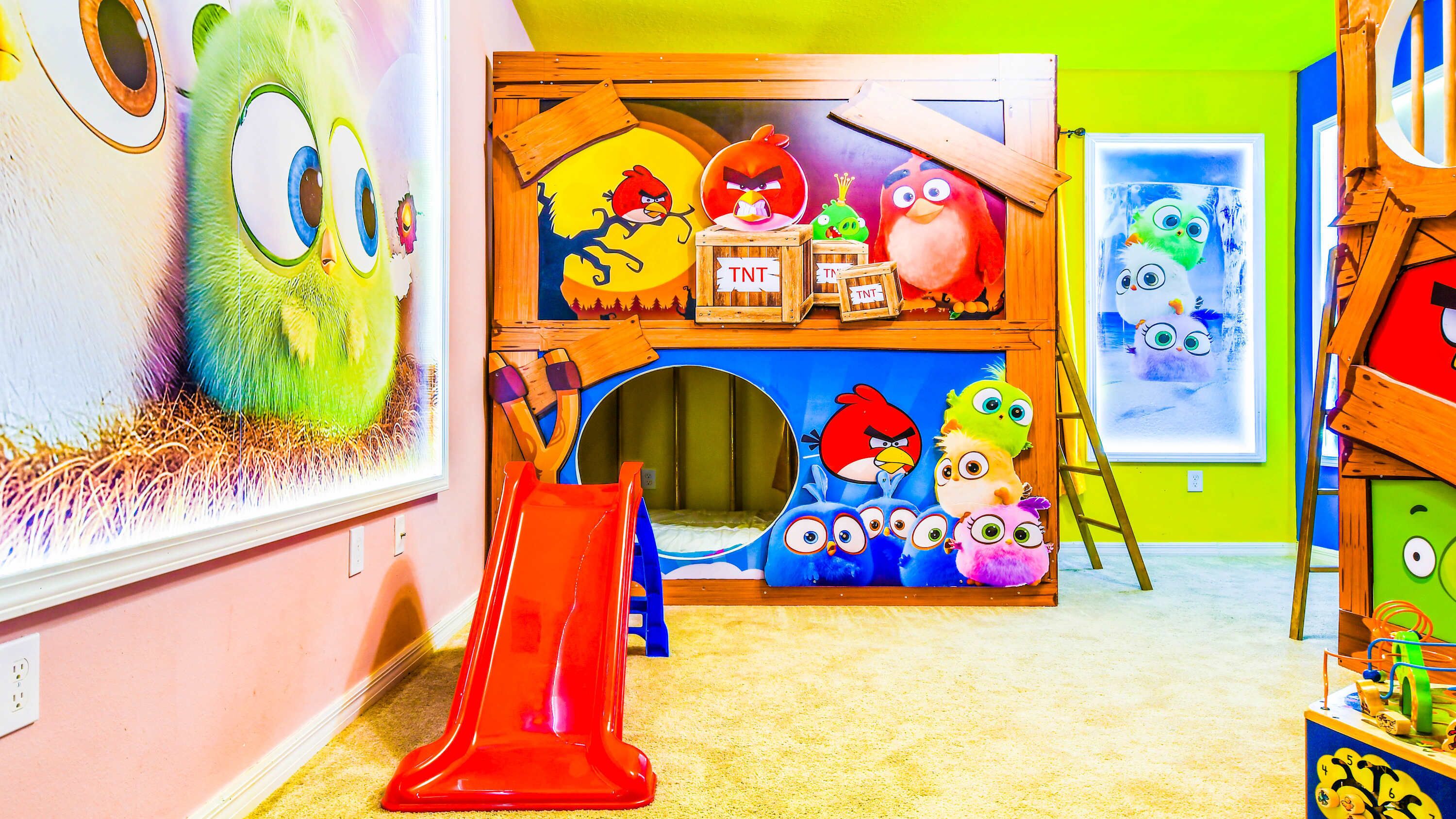 Une salle de jeux pour enfants avec un toboggan, un mur décoré d’un grand personnage de dessin animé et une étagère remplie de jouets Angry Birds.