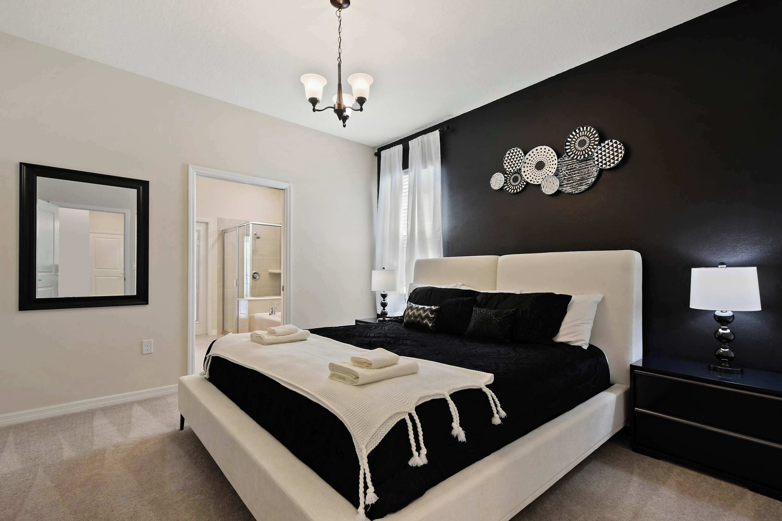 A bedroom with a large bed, a bedside table with a lamp, a mirror, and a wall art piece.
