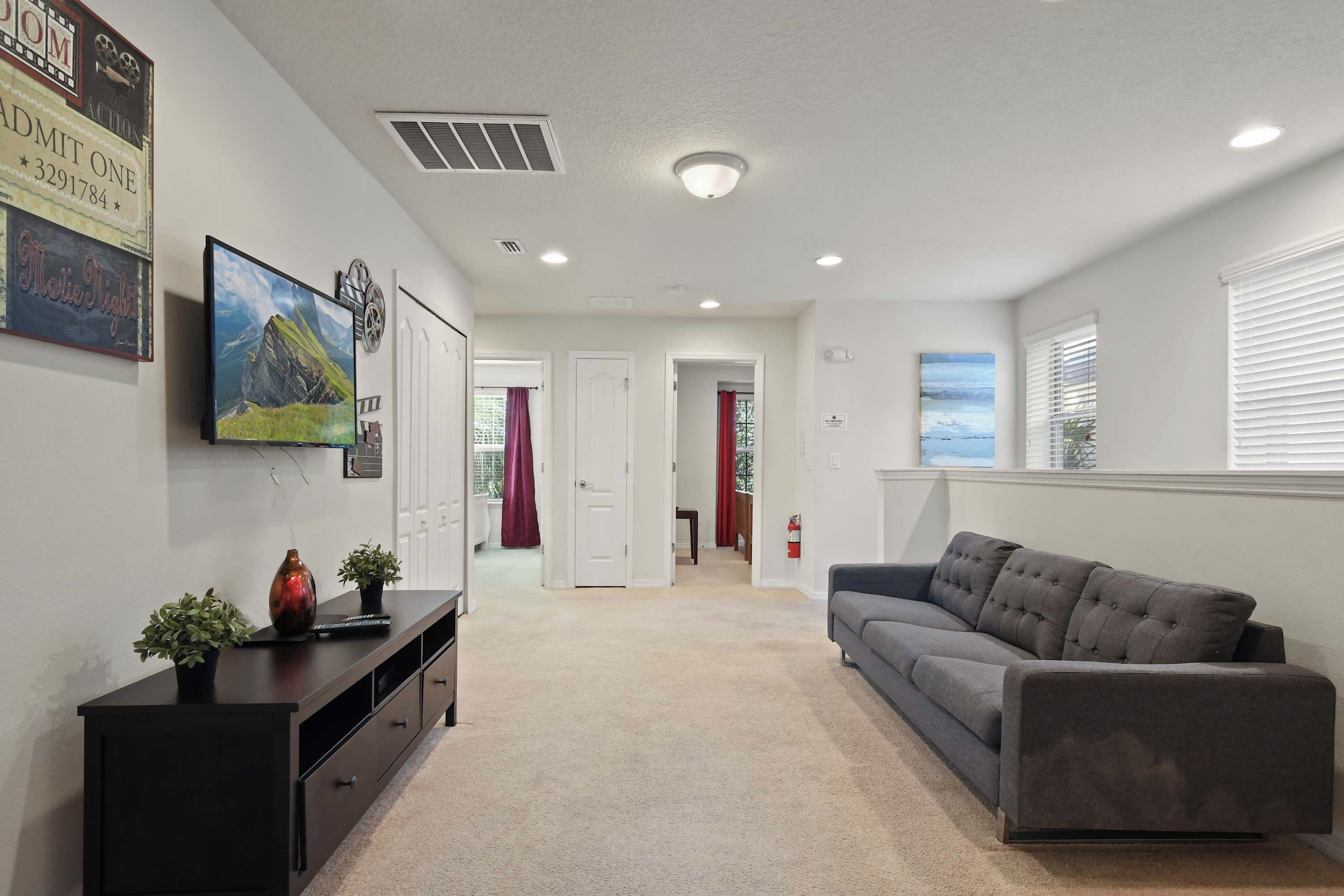 A living room with a grey sofa, a flat-screen TV mounted on the wall, and a small table with a vase and a plant.