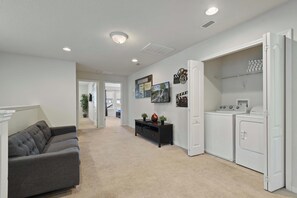 A modern living room with a grey sofa, a hallway leading to a bedroom, and a laundry area with a washing machine and dryer. - Free Waterpark Access! 5br Resort Townhome w/ Pool (Kissimmee)
