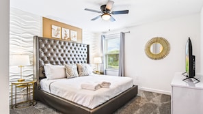 A modern bedroom with a tufted headboard, a large window with blinds, a ceiling fan, and a round mirror on the wall.