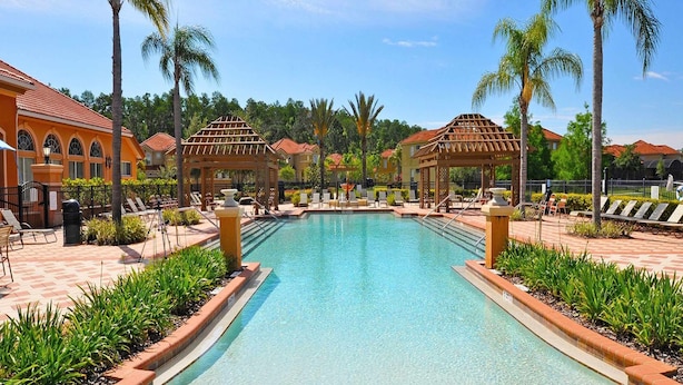 A swimming pool with lounge chairs and palm trees, surrounded by buildings with red-tiled roofs. - Family-friendly Luxury Home Near Disney & More (Kissimmee)