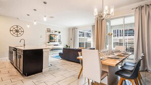 A modern kitchen with a central island, a dining area with a wooden table and chairs, and a large window with curtains. - 4BR Retreat in Champions Gate w/ Private Pool (Davenport)