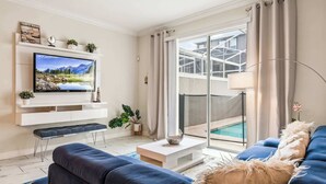 A modern living room with a flat-screen TV, a sofa, a coffee table, and a view of a swimming pool through large glass doors. - 4BR Retreat in Champions Gate w/ Private Pool (Davenport)