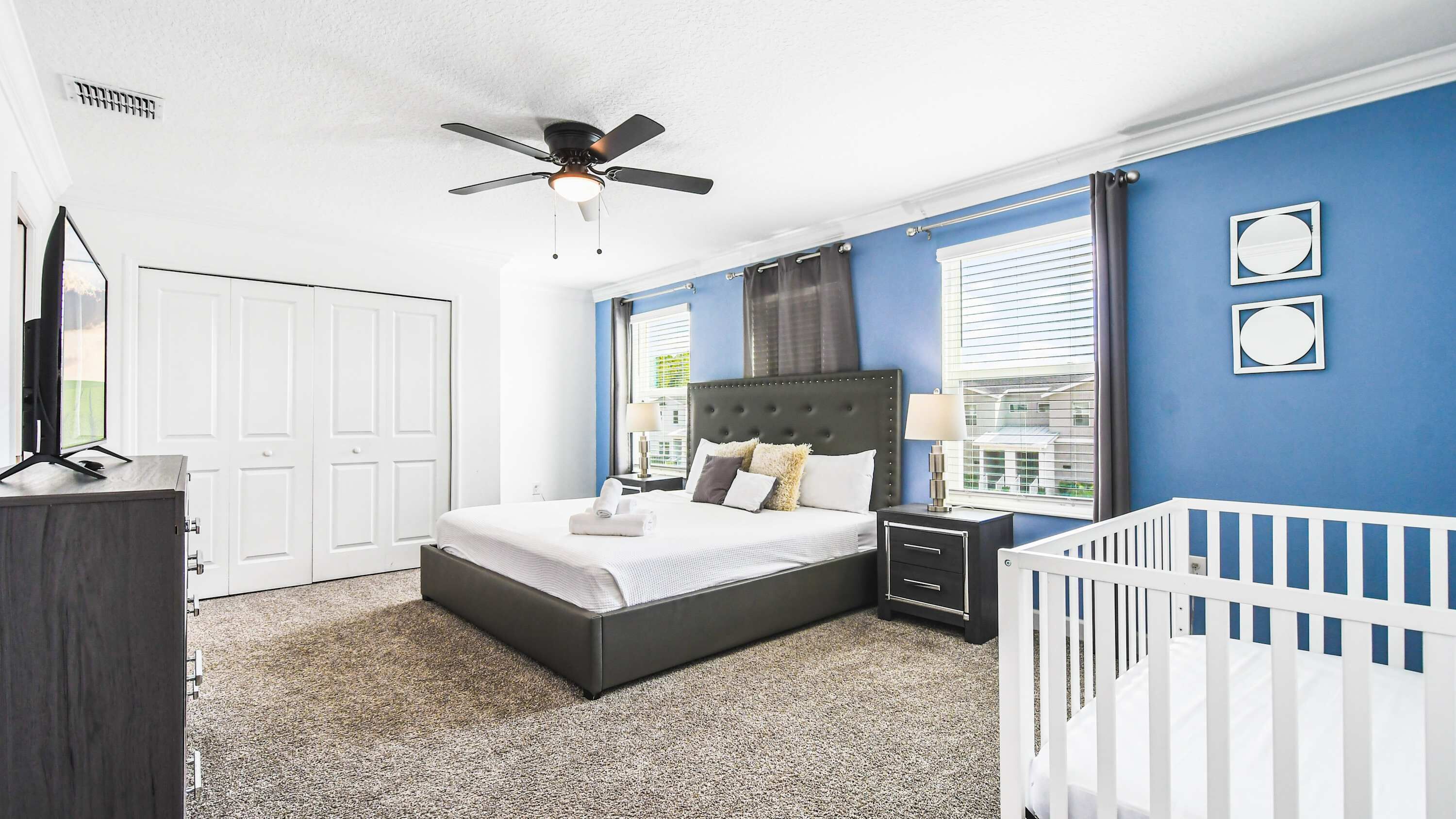A bedroom with a bed, a nightstand, a crib, a ceiling fan, and a window with blinds.