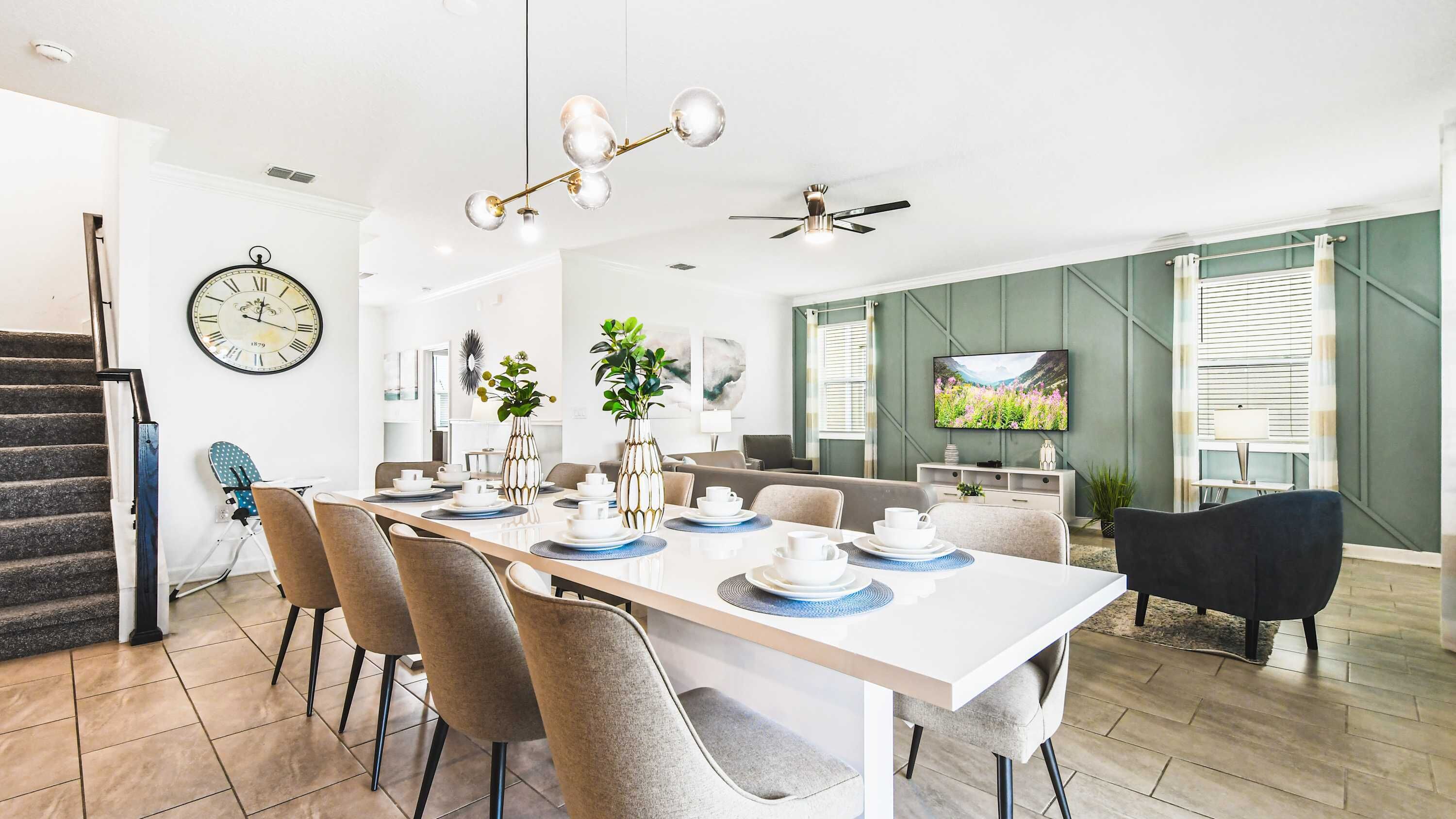 A modern dining area with a white table set for six, surrounded by grey chairs. A large wall clock and a flat-screen TV are visible in the background.