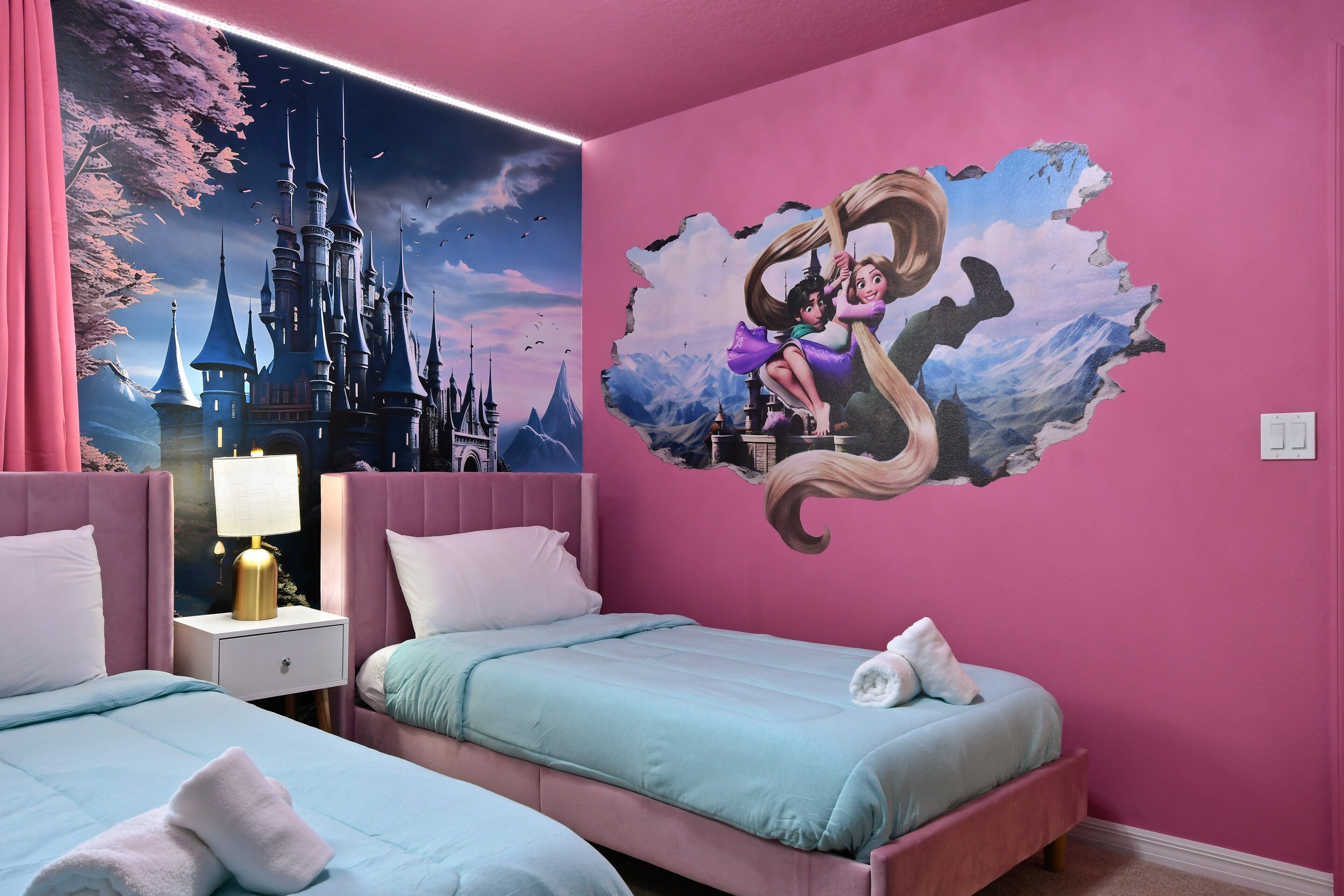 Un dormitorio con temática rosa, con dos camas, una mesita de noche con lámpara y un gran mural de una princesa en la pared.