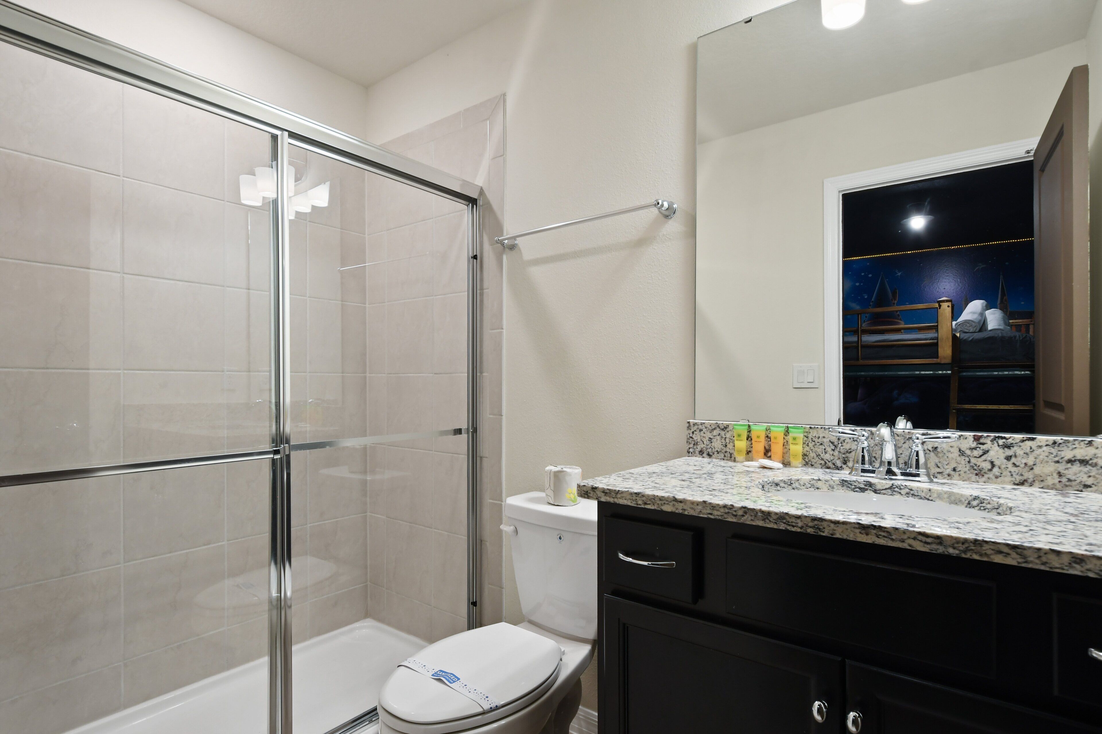 A bathroom with a glass shower enclosure, a toilet, a sink with a granite countertop, and a mirror with a night sky scene.