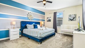 Quarto com cama estofada azul, duas mesas de cabeceira com abajures, um berço branco e um ventilador de teto.