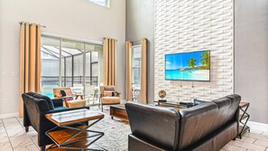A modern living room with a large TV, leather sofas, a coffee table, and a glass door leading to a balcony. - Storey Lake Cozy Resort Home Pool Spa Disney Theme (Kissimmee)