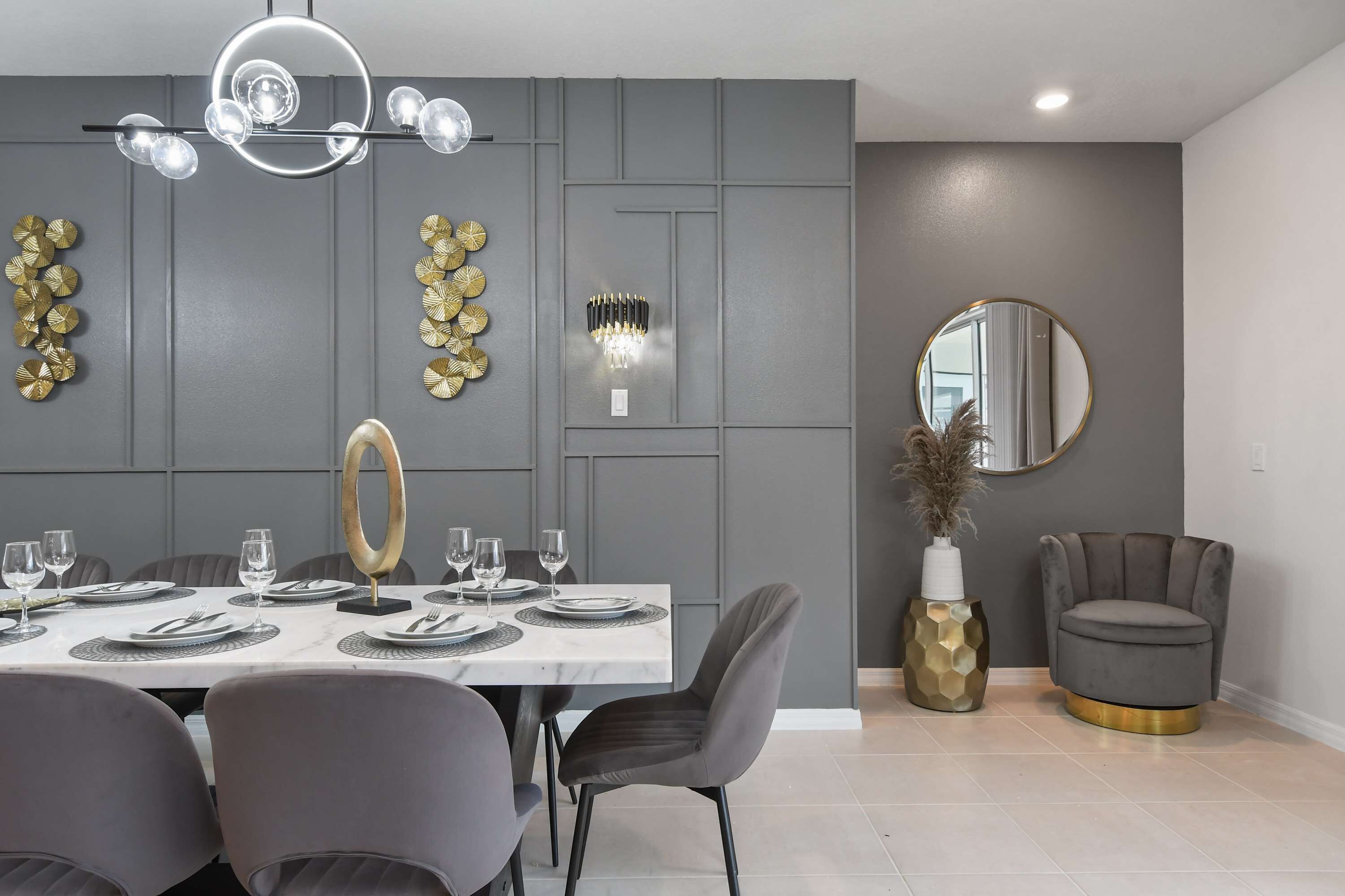 Un coin repas avec une table blanche dressée pour quatre personnes, des chaises à revêtement gris, un lustre, des assiettes décoratives accrochées au mur et un miroir rond.