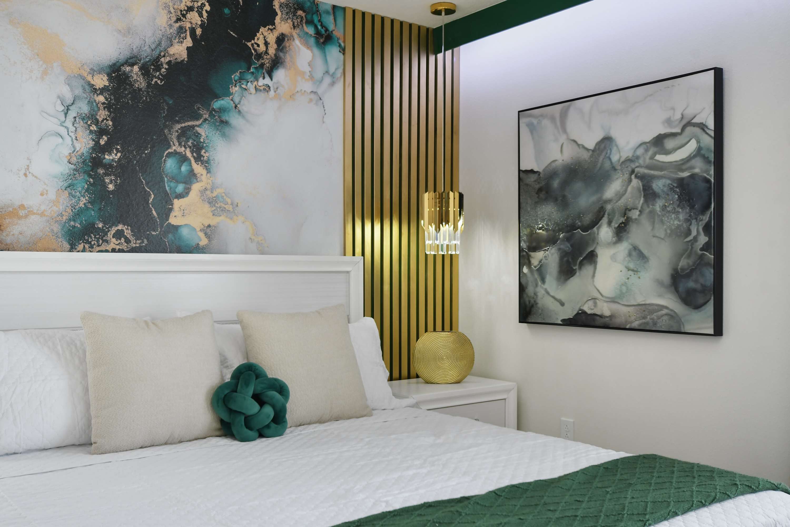 Een slaapkamer met een bed, kussens, een nachtkastje, een lamp en abstracte wanddecoratie.