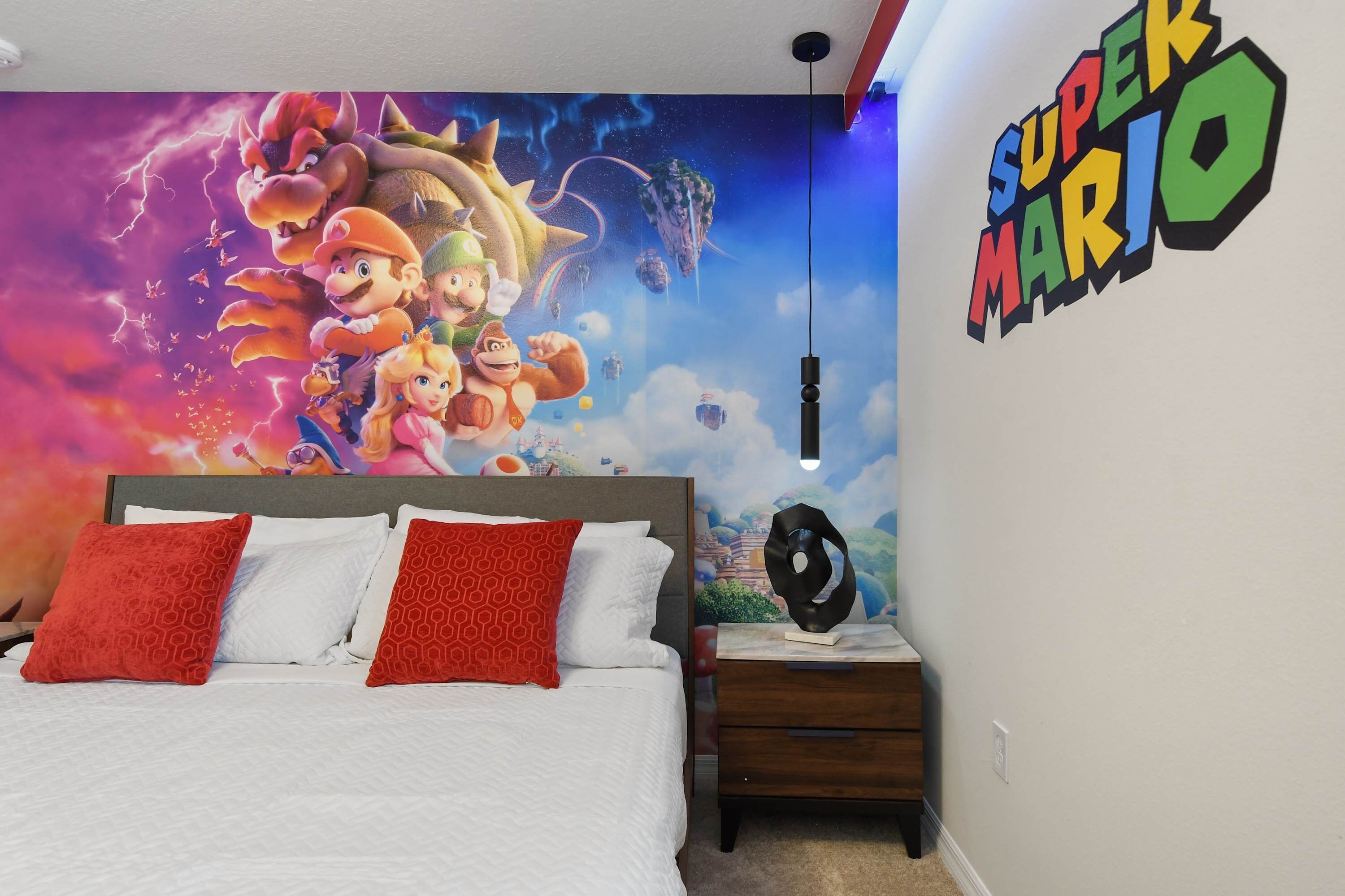 Un dormitorio con un mural temático de Super Mario, una cama con ropa de cama blanca y roja, y una mesita de noche con una lámpara.