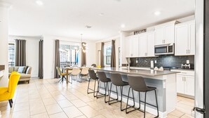 Una cocina moderna con armarios blancos, una isla central con taburetes de bar, un comedor con una silla amarilla y vistas de la ciudad a través de amplias ventanas.