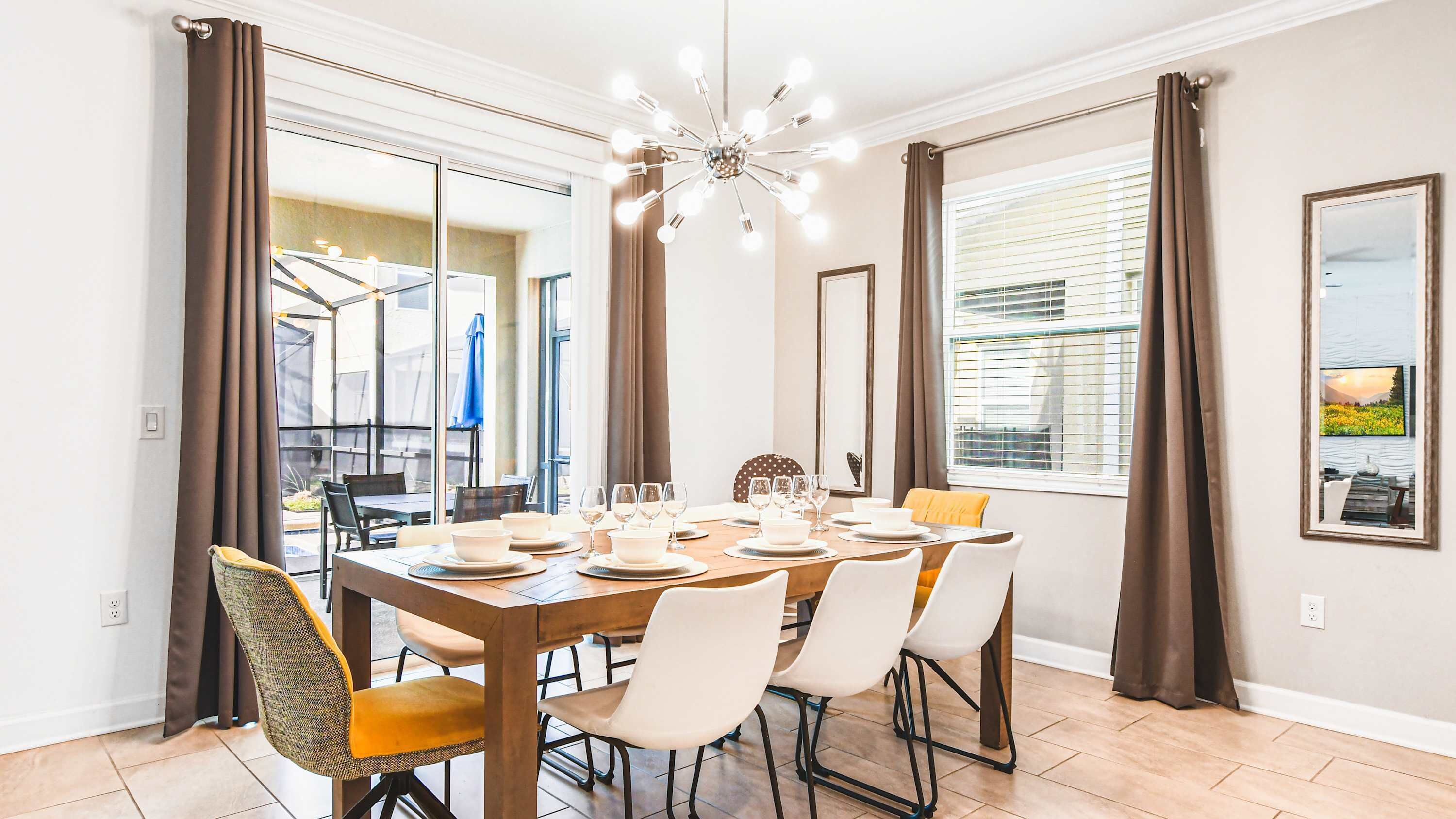 A dining area with a wooden table set for six, surrounded by chairs, with a modern chandelier above.