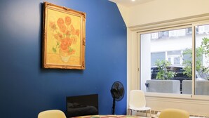 A room with a blue wall, a painting of flowers, a round table with a colorful tablecloth, a fan, and a window with a view of the outside. - Colorful 1BR in Beaugrenelle - 5min to Metro (Paris)
