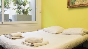 A neatly made bed with white linens, a window with a view of greenery, and a yellow wall. - Colorful 1BR in Beaugrenelle - 5min to Metro (Paris)