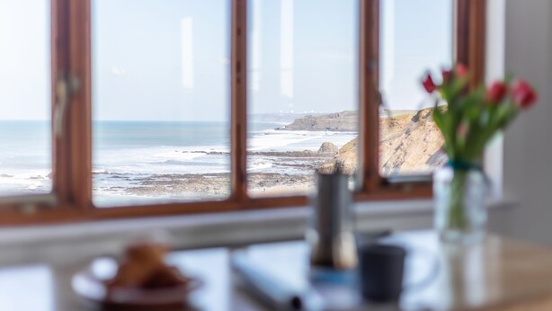 Uitzicht op de oceaan door een raam, een vaas met bloemen en een bord eten op een tafel.