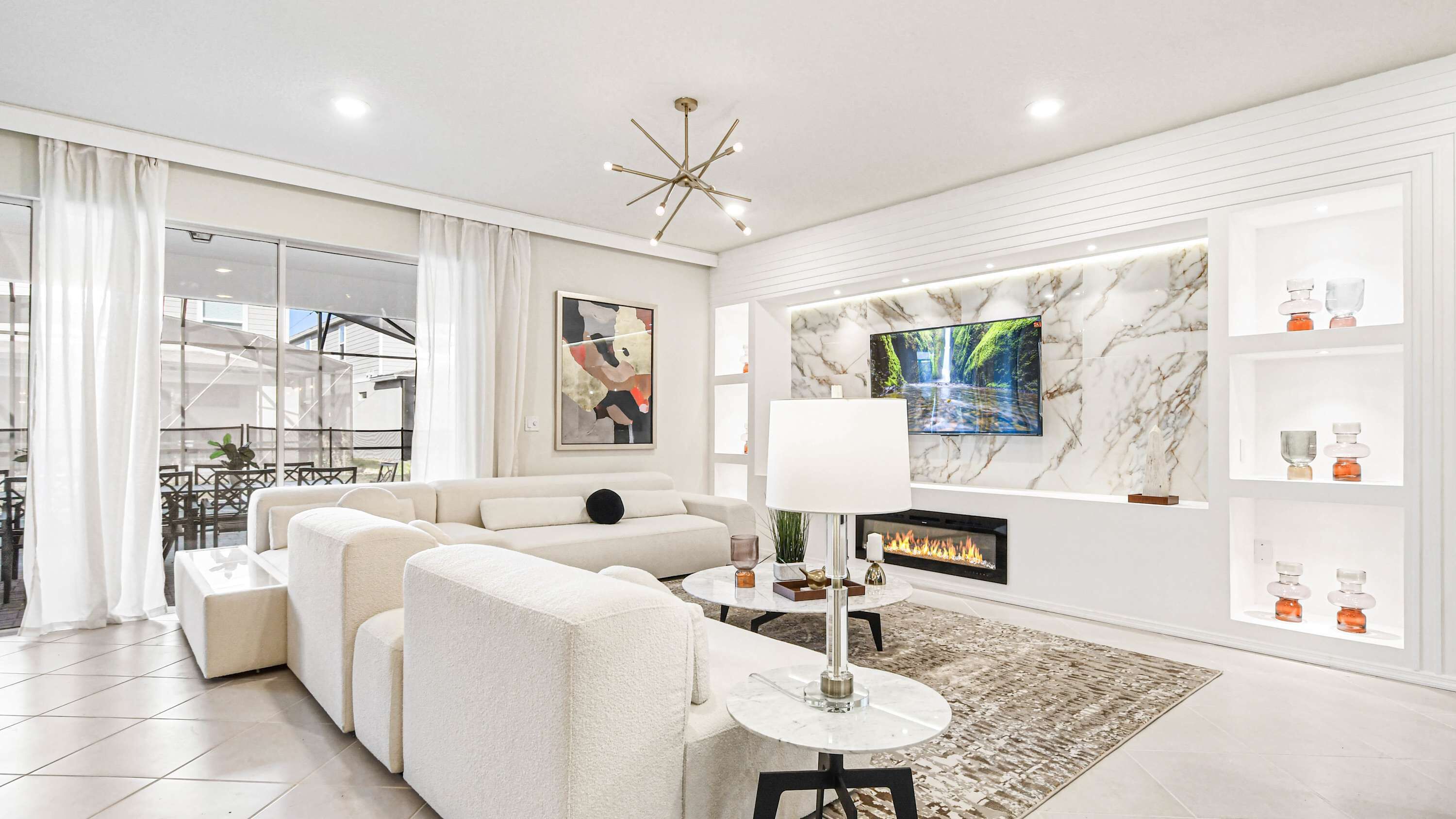 A modern living room with a sectional sofa, a fireplace, a TV mounted on a marble wall, and a glass-enclosed balcony.