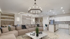 Una sala de estar moderna con un sofá seccional, una mesa de centro y una cocina con alacenas blancas y un refrigerador de acero inoxidable.