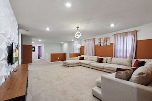 A modern living room with a sectional sofa, a wooden coffee table, and a flat-screen TV mounted on the wall. - 7BR Solterra Resort - Newly Renovated Home w Pool (Davenport)