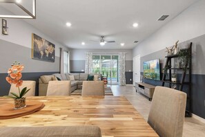 A spacious living room with a wooden dining table, sofas, and a TV on the wall. - 7BR Solterra Resort - Newly Renovated Home w Pool (Davenport)