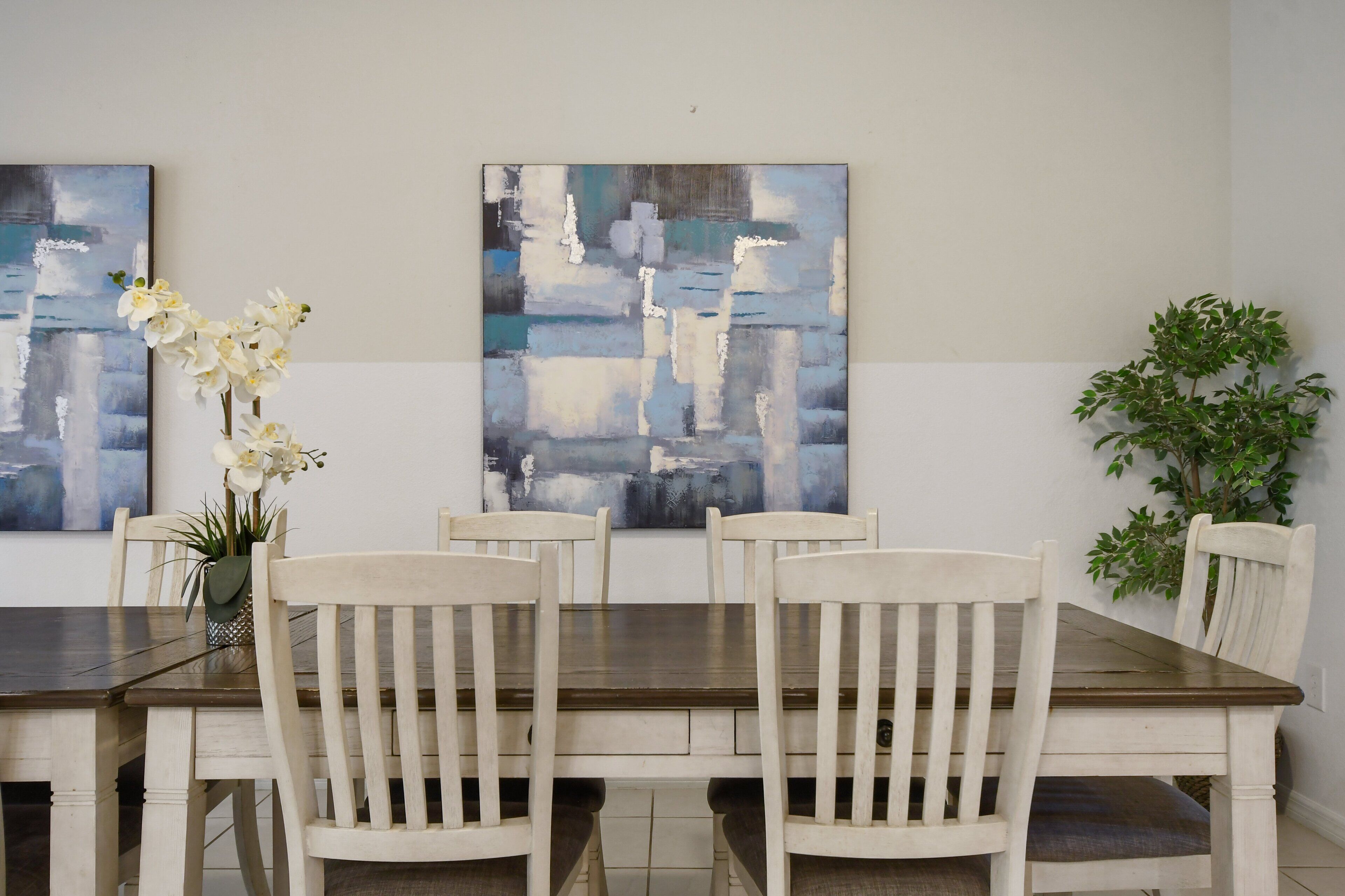 A dining area with a wooden table and six white chairs, flanked by two abstract paintings and a potted plant.