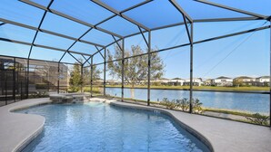 A swimming pool with a glass enclosure, surrounded by a landscaped area and a view of houses across a body of water. - Elegant Home With Pool & Spa, Close to Disney (Kissimmee)