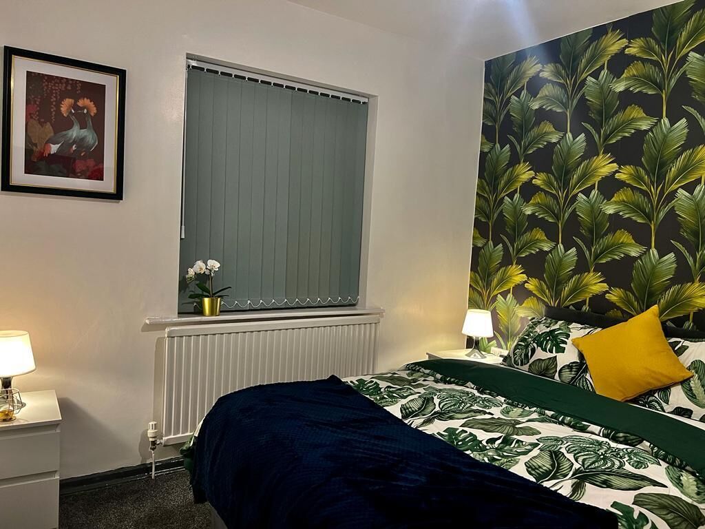 Une chambre avec un lit, une fenêtre avec des stores, un radiateur, une table de chevet avec une lampe, un tableau encadré au mur et un grand mur végétal.
