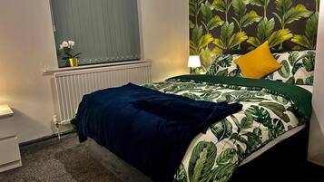 A bedroom with a bed, a window with a curtain, a radiator, a lamp, and a wall with a leafy pattern.