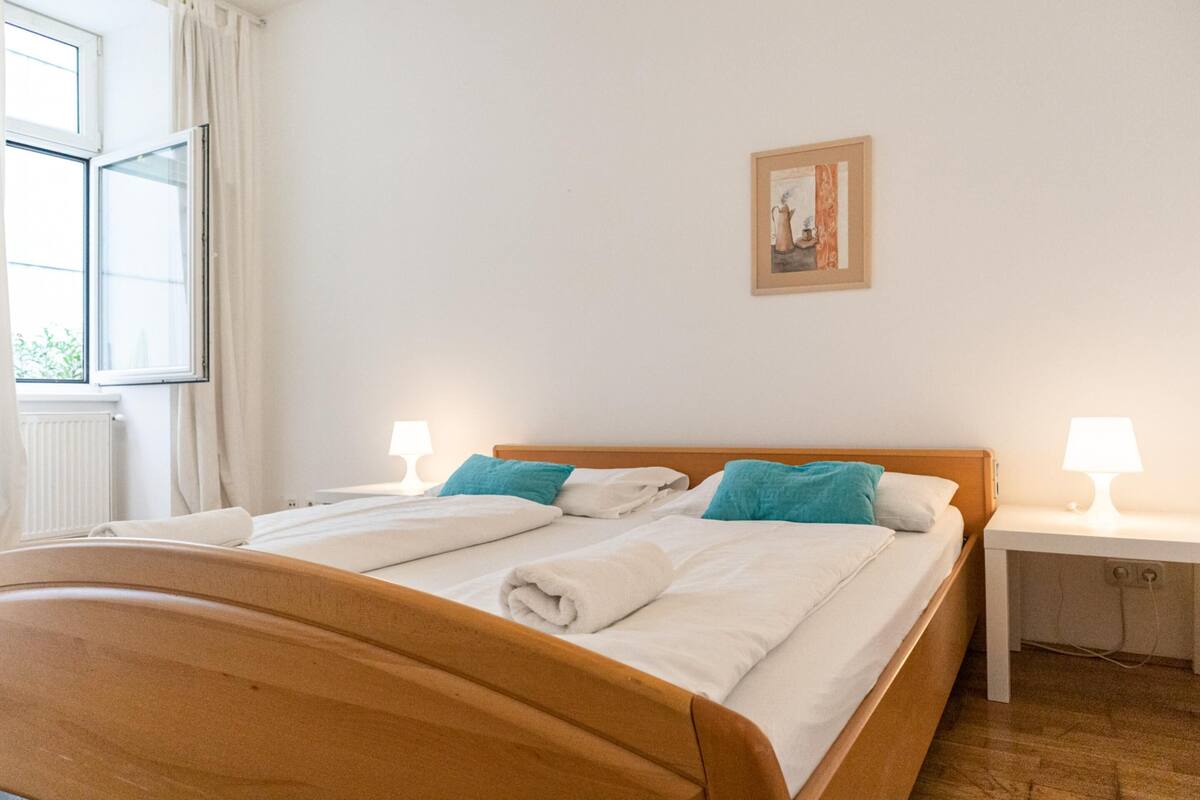 A neatly made bed with white linens and blue pillows, a bedside table with a lamp, and a framed picture on the wall.