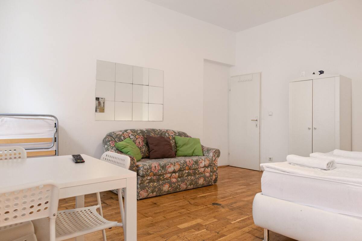 A room with a wooden floor, a white sofa with floral upholstery, a white table with a remote control, and a white wardrobe.