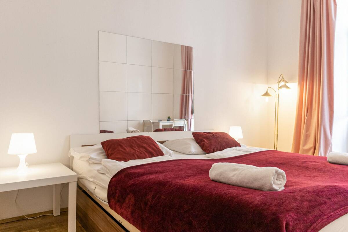 A neatly made bed with a red blanket, a white bedside table with a lamp, a large mirror, and a floor lamp.