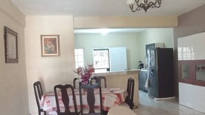 Un coin repas avec une table dressée pour quatre personnes, des chaises, un lustre, un réfrigérateur et, au fond, une cuisine.