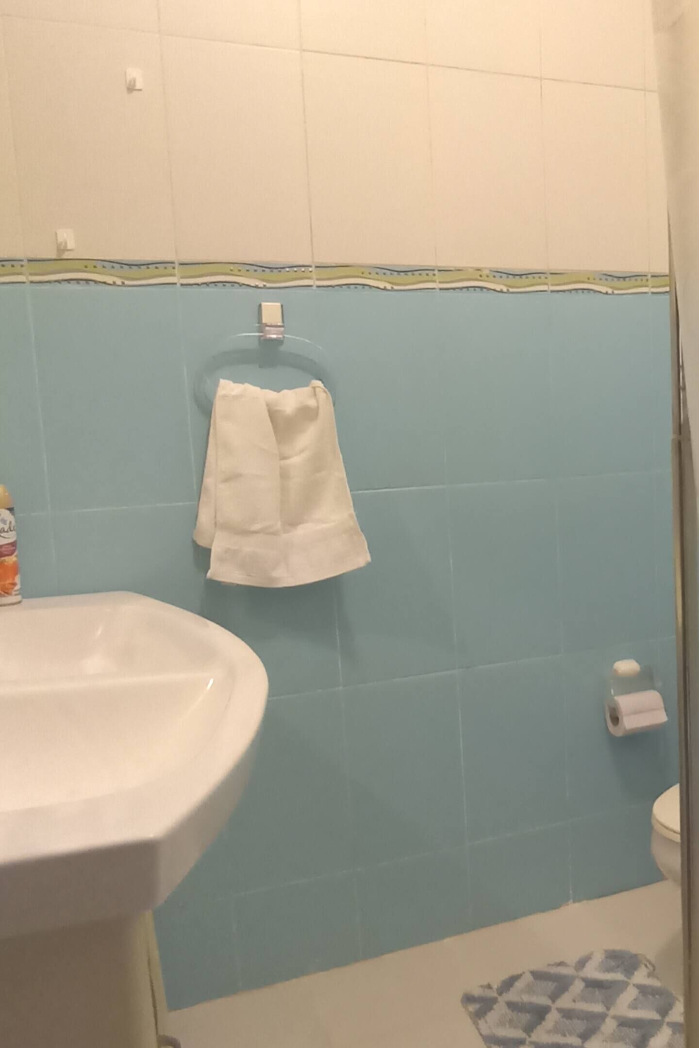 A bathroom with a white sink, blue tiled walls, a white towel hanging on a rack, and a toilet with a paper towel holder.