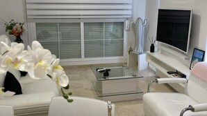 A modern living room with a white sofa, a glass coffee table, a flat-screen TV, and a large window with blinds. - Magnifique, et Luxueux Appartement en Top Résidence à Benalmadena (Benalmádena)