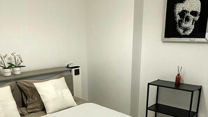 A neatly made bed with a white textured bedspread, a headboard, and two white pillows. There is a small black nightstand with a red vase and a framed skull artwork on the wall. - Magnifique, et Luxueux Appartement en Top Résidence à Benalmadena (Benalmádena)