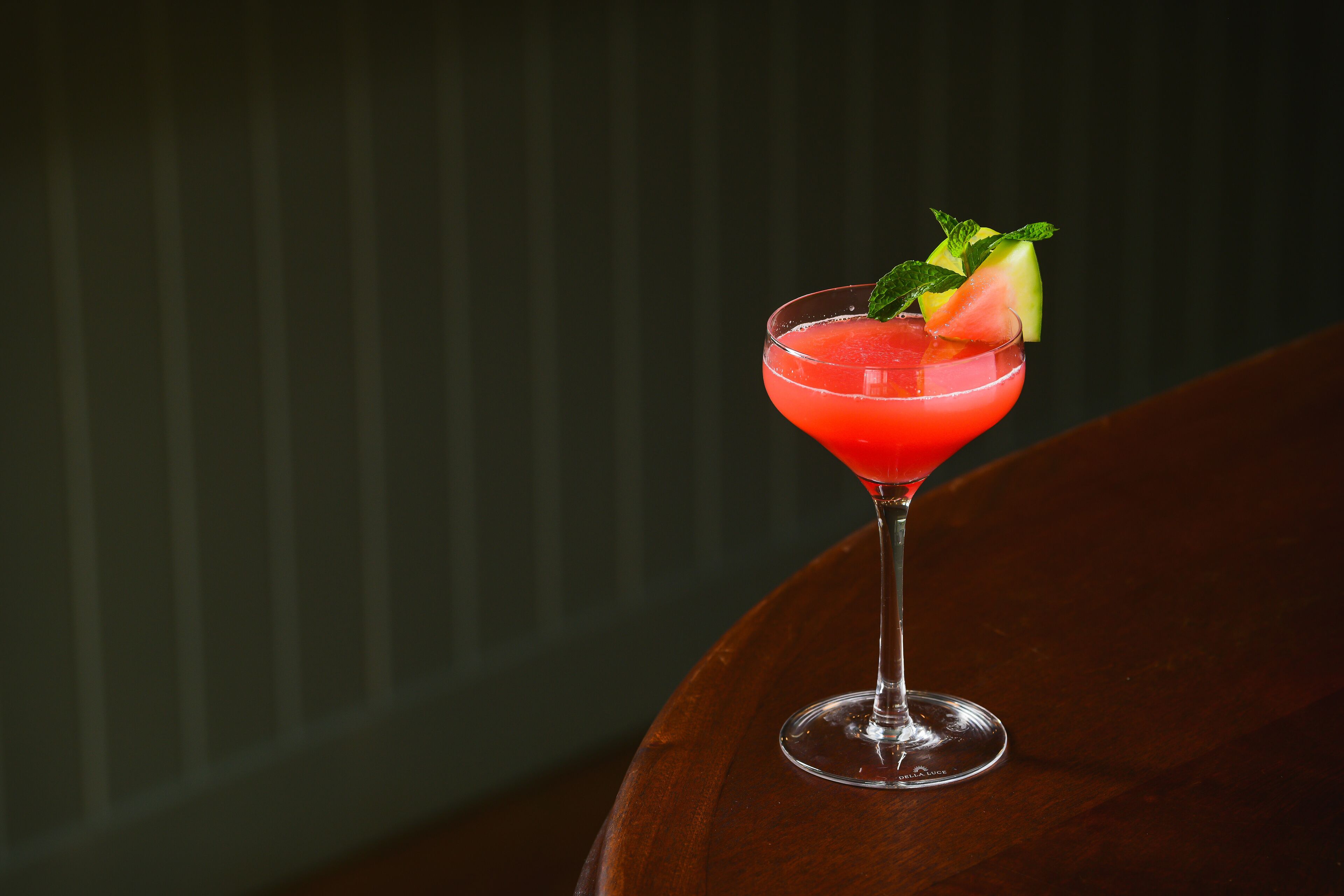 Un cóctel en copa de martini adornado con una hoja de menta y una rodaja de fruta sobre una mesa de madera.