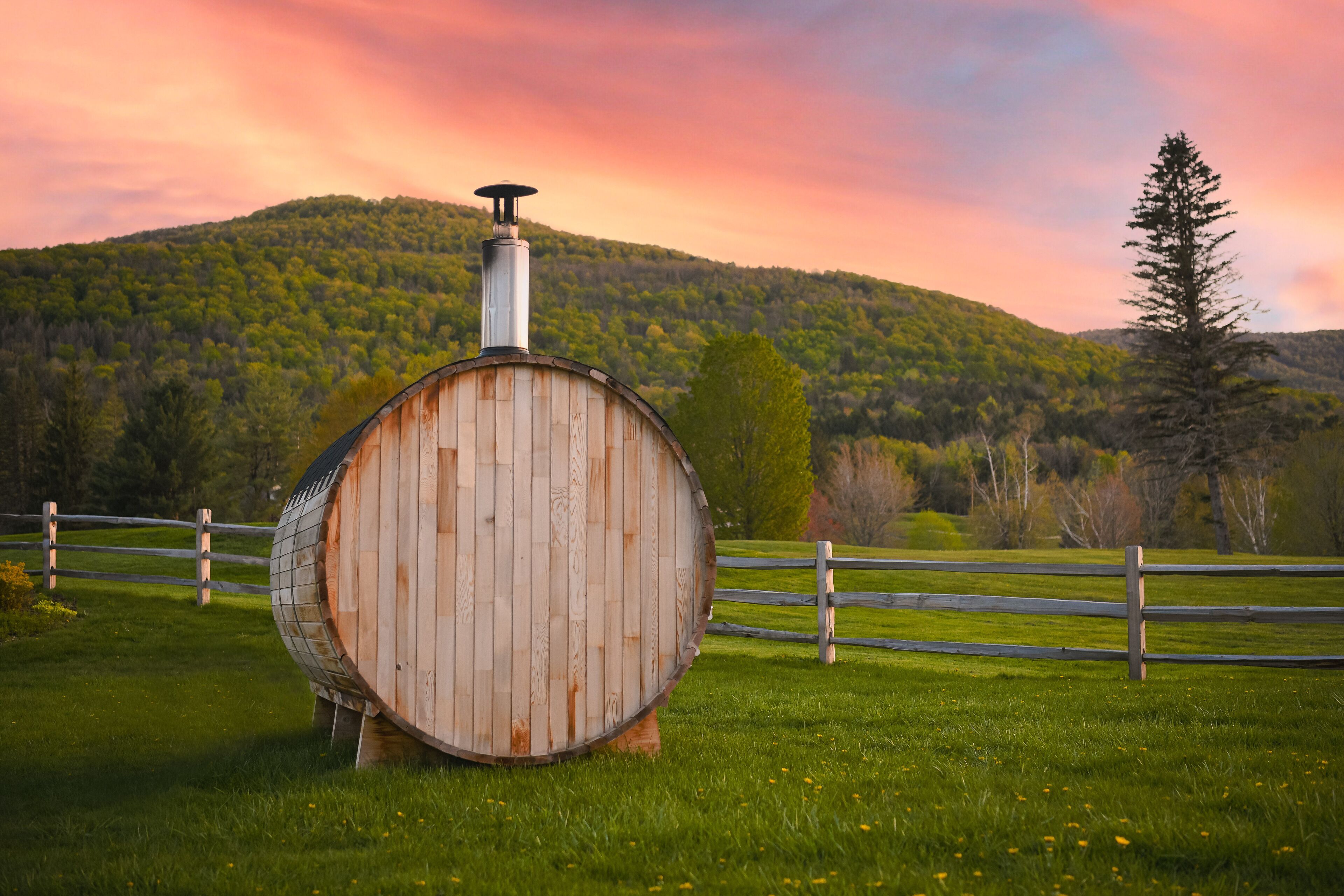 A rustic wooden barrel with a chimney, set in a grassy field with a wooden fence and a forested hill in the background.