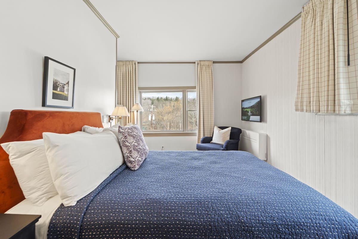 A bedroom with a large bed, a nightstand, a chair, a window with curtains, a framed picture on the wall, and a TV mounted on the wall.