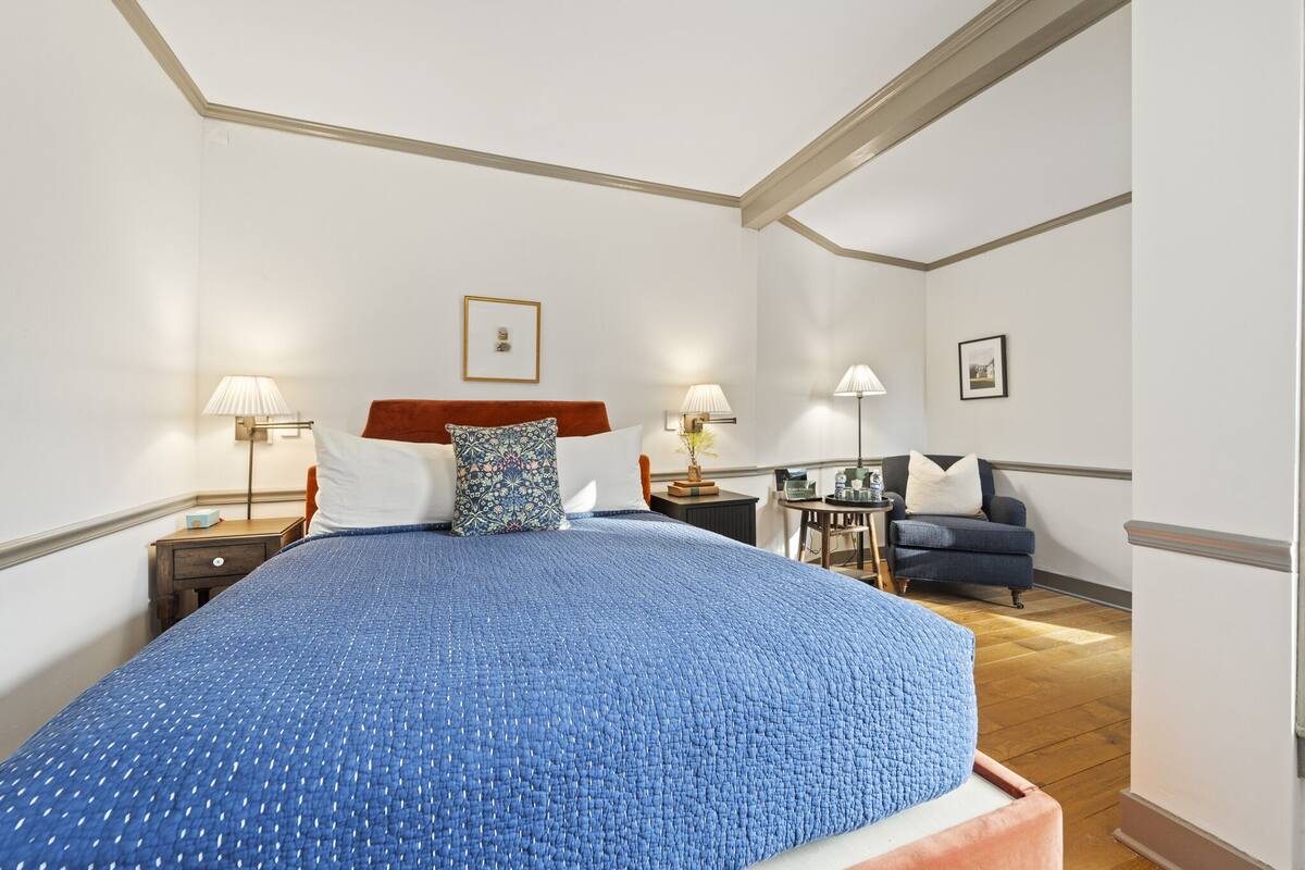 A spacious bedroom with a large bed, two bedside tables with lamps, a small side table, a chair, and a framed picture on the wall.